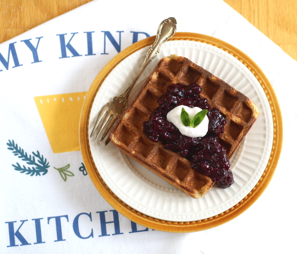 Plate with a waffle and fruit compote over a tea towel with "my kind kitchen" on it.